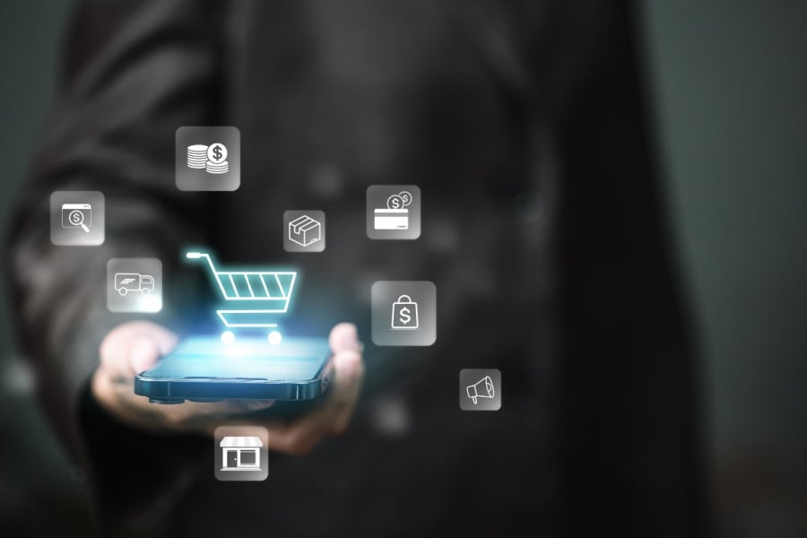 Person presents smartphone with glowing shopping cart and retail icons, representing mobile e-commerce, online payments, checkout, marketing automation and omnichannel sales growth